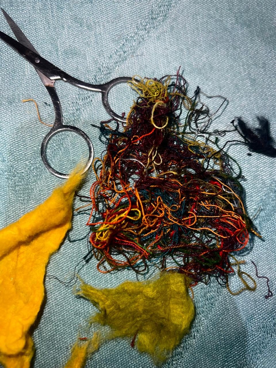 Embroidery scissors beside a nest of cut thread ends in yellow, rust, teal and red, with raw dyed cotton fluff.